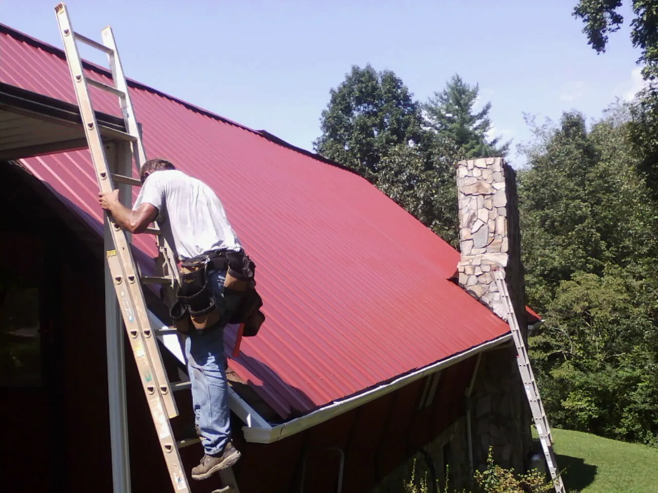Roofer on a ladder beside a red metal roof and stone chimney — Soffit & Fascia Repair by Roofing Brothers in Pope Army Airfield